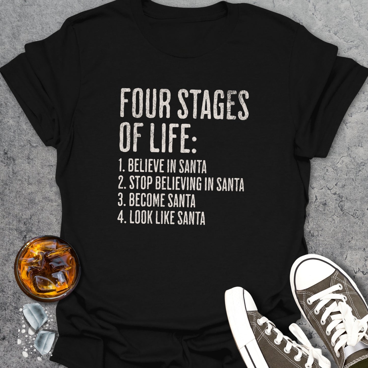 Black t-shirt with 'Four Stages of Life' text on a concrete surface with a glass of whiskey and sneakers.