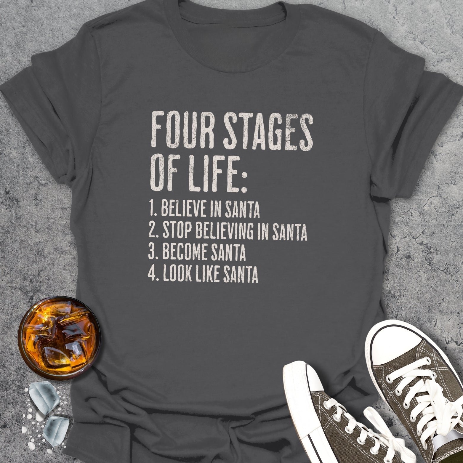 Gray t-shirt with 'Four Stages of Life' text, placed on a textured surface with a glass of whiskey and sneakers.